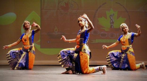 Three girls dressed in purple, orange and gold Bharatanatyam dance outfits perform on stage.